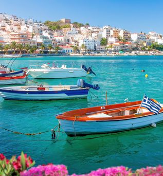 Mehrere kleine Boote mit griechischer Flagge liegen an einer Hafenmauer, im Hintergrund Häuser an einem Hang und blühende pinke Blumen im Vordergrund.