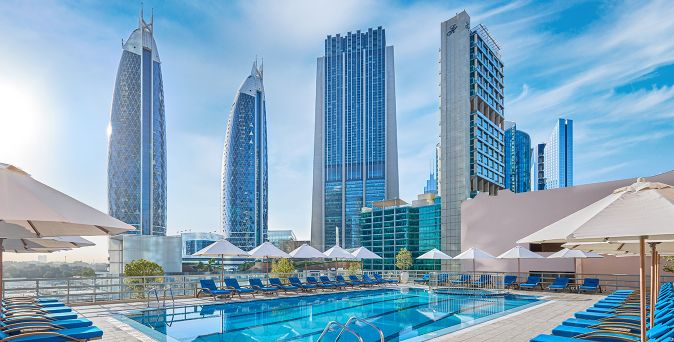 Außenpool mit Sonnenliegen und Sonnenschirmen vor den markanten Hochhäusern der Dubai-Skyline am Rose Rayhaan by Rotana.