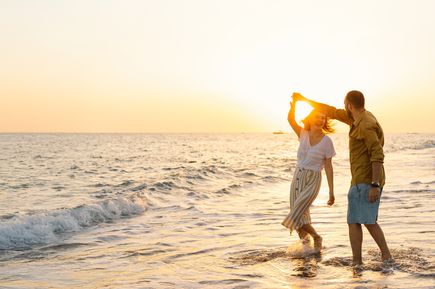 Paar tanzt am Strand im flachen Wasser bei Sonnenuntergang. Frau trägt gestreiften Rock und weißes T-Shirt, Mann Shorts und Hemd.
