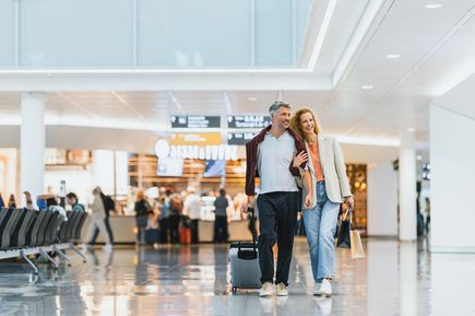 Zwei Reisende mit Rollkoffer und Einkaufstüten im Terminal des Flughafens München, moderne Abflughalle mit Shops und Sitzbereichen im Hintergrund