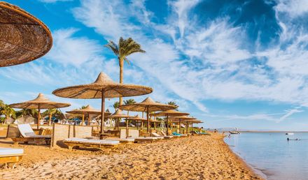 Strand am Roten Meer in Ägypten mit strohbedeckten Sonnenschirmen, Liegen und ruhigem, flach abfallendem Wasser unter blauem Himmel
