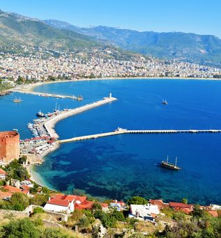 Luftaufnahme des Hafens von Alanya in der Türkei mit geschwungenem Wellenbrecher, mehreren Booten im türkisblauen Meer