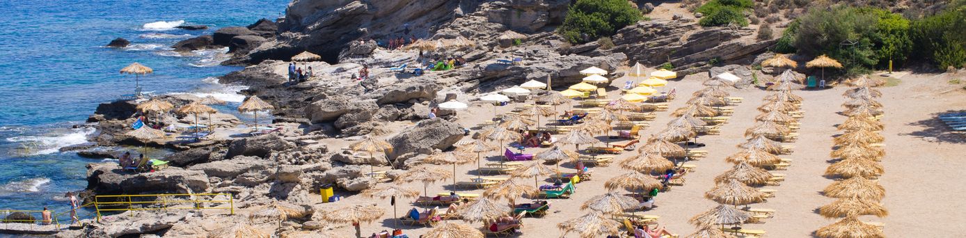 Felsige Küste mit mehreren Sonnenschirmen und Liegen, Menschen entspannen am Meer bei klarem Himmel auf Rhodos.