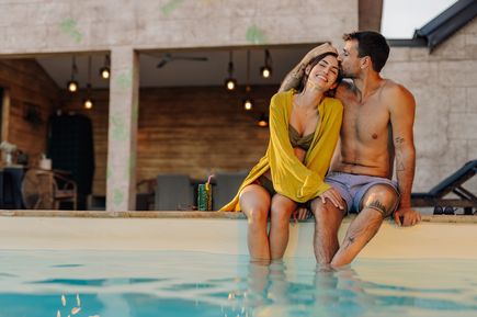 Junges Paar sitzt am Poolrand, lächelt und zeigt Zuneigung, mit sommerlicher Atmosphäre im Hintergrund