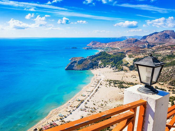 Blick von einer Terrasse mit Holzgeländer und Laterne auf den Tsambika-Strand mit türkisblauem Meer und bewölktem Himmel auf Rhodos.
