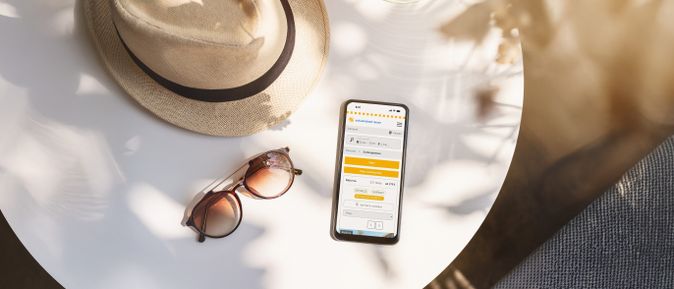 Sommerliche Szene mit Sonnenhut, Sonnenbrille und Smartphone mit Webseite von schauinsland-reisen auf einem weißen Tisch