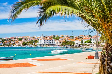 Küstenstadt Novalja auf der Insel Pag in Kroatien mit türkisblauem Meer, Hafenpromenade, kleinen Booten und Palmen im Vordergrund