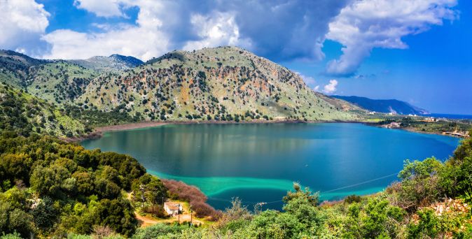 Blick auf den Kournas-See in Kreta, umgeben von grünen Hügeln und Bergen unter einem bewölkten Himmel.