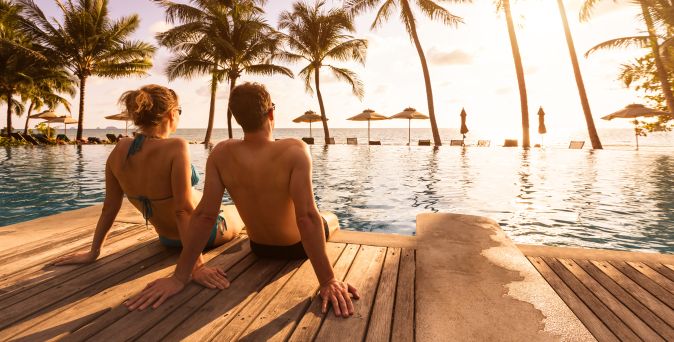 Paar sitzt am Poolrand mit Blick auf Palmen und Meer bei Sonnenuntergang