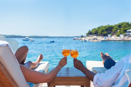 Zwei Personen stoßen mit Sommerdrinks an, während sie auf Liegen direkt am klaren Meer entspannen