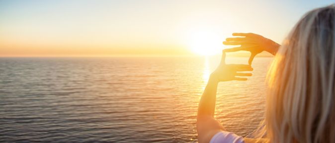 Person betrachtet den Sonnenuntergang über dem ruhigen Meer und formt mit den Händen einen Rahmen um die Sonne