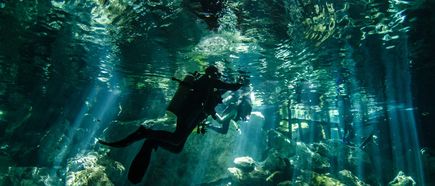 Zwei Taucher in voller Ausrüstung schwimmen unter Wasser über felsigem Grund, Sonnenstrahlen durchdringen die Wasseroberfläche.