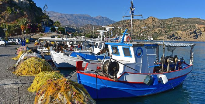 Fischerboote mit blauen Rümpfen liegen an einem Hafen, im Vordergrund liegen gelbe Fischernetze auf dem Kai, im Hintergrund Hügel und ein Gebäude.