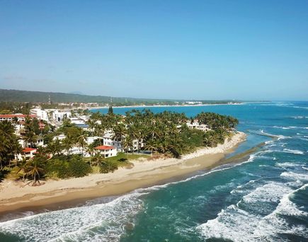 Küste mit Sandstrand, Wellen und mehreren weißen Gebäuden hinter Palmen unter blauem Himmel.
