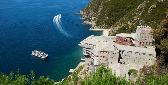 Blick von oben auf ein großes Klostergebäude an einer steilen Küste, davor ein Ausflugsboot auf blauem Wasser mit sichtbarer Bugwelle.