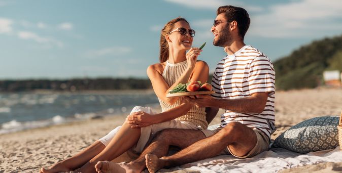 Paar sitzt bei einem Picknick am Strand, lacht gemeinsam und genießt frisches Obst mit Blick auf das Meer