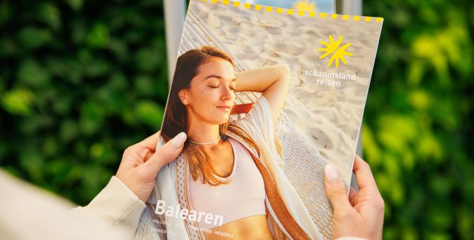 Person hält Katalog mit Titel 'Balearen' und Logo von 'Schaunisland reisen', auf dem eine Frau in weißem Top am Strand liegt.