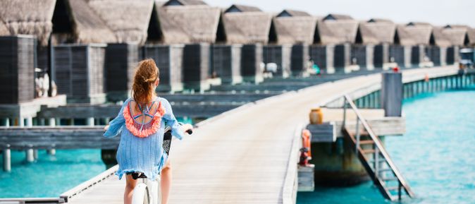 Frau mit Fahrrad auf einem Steg vor Wasserbungalows über klarem Wasser