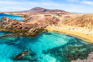 Klares türkisblaues Wasser an einer felsigen Küste mit goldgelbem Sandstrand und Bergen im Hintergrund unter blauem Himmel.