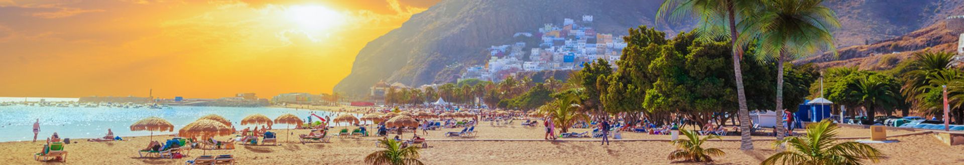 Strand mit Sonnenschirmen und Palmen vor Bergen bei Sonnenuntergang mit Wolken am Himmel