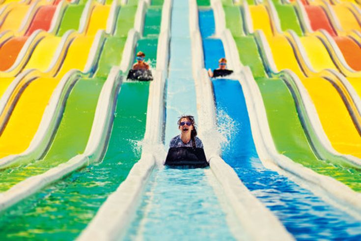 Kinder rutschen auf bunten, nebeneinander angeordneten Wasserrutschen mit Matten in einem Wasserpark, kräftige Farben wie Gelb, Grün und Blau