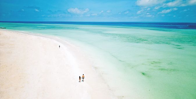 Weitläufiger, heller Sandstrand mit türkisfarbenem, flachem Meer; zwei Personen gehen entlang der Küstenlinie unter blauem Himmel.
