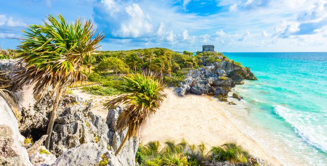 Küste von Tulum in Mexiko mit Sandstrand, türkisfarbenem Meer und Ruinen auf einer Klippe unter blauem Himmel