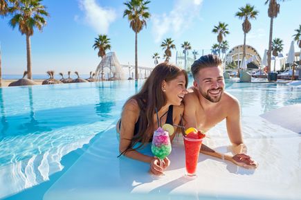 Zwei Erwachsene entspannen nebeneinander im flachen Pool mit bunten Cocktails, umgeben von Palmen und Strandkulisse