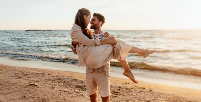 Mann trägt Frau in hellem Kleid am Strand vor ruhigem Meer bei Sonnenuntergang