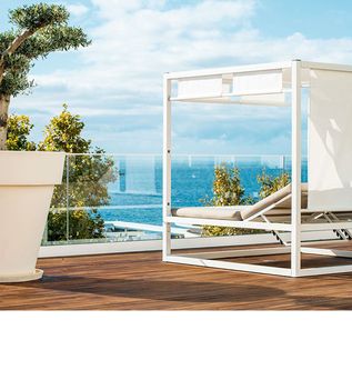 Weißes, modernes Tagesbett mit Stoffdach auf Holzterrasse, daneben großer weißer Blumentopf mit Baum, Meer im Hintergrund.