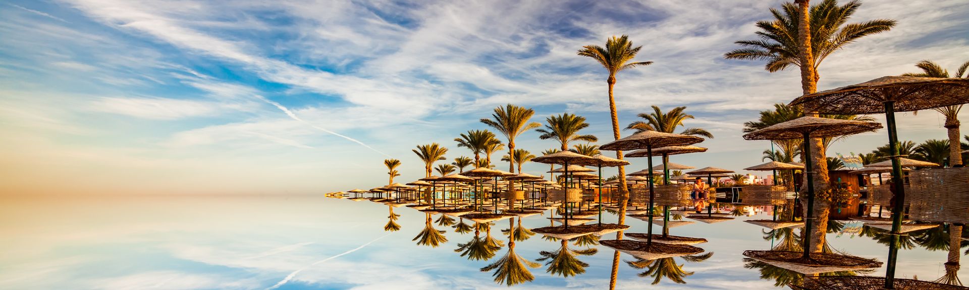 Spiegelglatte Wasseroberfläche mit mehreren Palmen und Strohdächern, die sich darin spiegeln, unter blauem Himmel mit Wolken.
