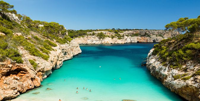 Klares türkisfarbenes Wasser in einer von Felsen und Pinien umgebenen Bucht auf den Balearen mit schwimmenden Menschen