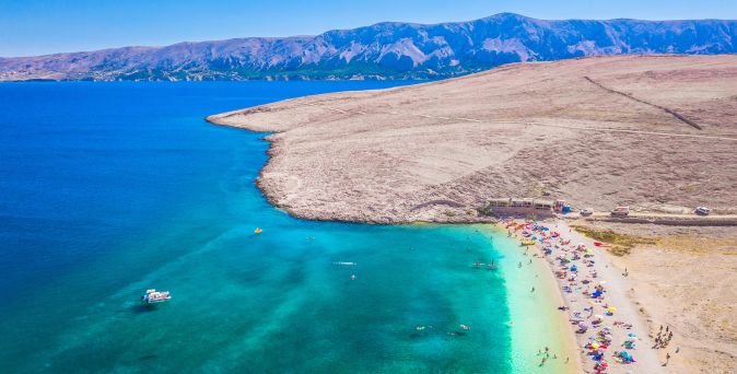 Luftaufnahme des Ručica-Strands auf der Insel Pag in Kroatien mit hellem Kiesstrand, türkisblauem Meer, Badegästen und karger Küstenlandschaft
