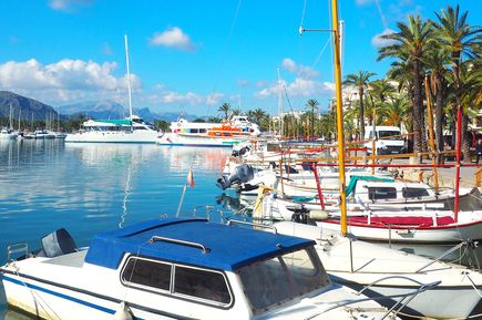 Boote und Yachten im Hafen von Alcúdia auf Mallorca, gesäumt von Palmen und Restaurants, mit Bergen im Hintergrund