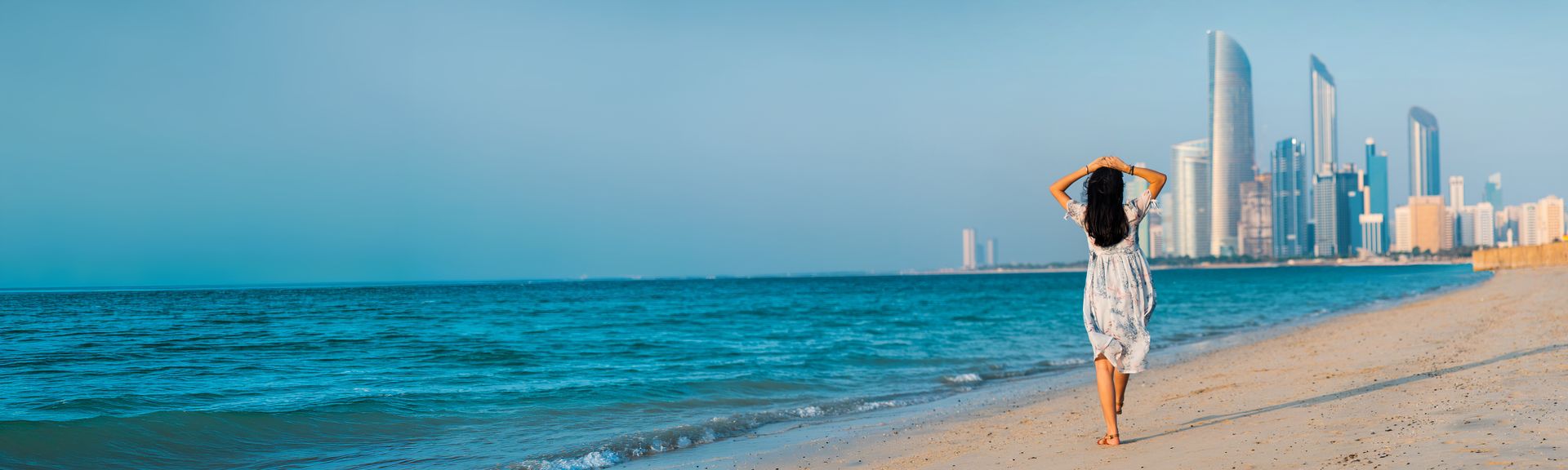 Frau mit langem Kleid steht am Strand und blickt auf die Skyline von Abu Dhabi