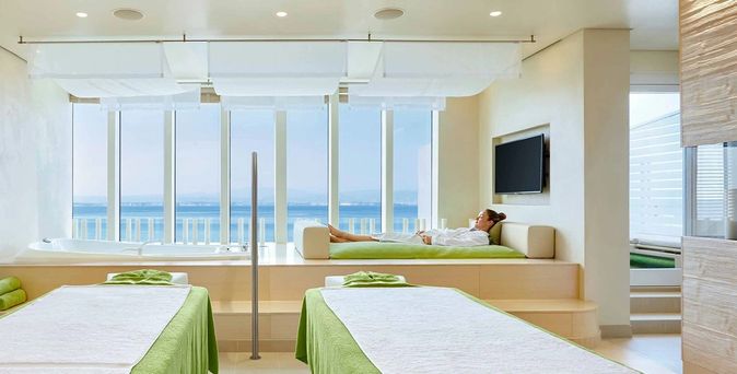 Modernes Spa mit zwei Massageliegen, großen Fenstern mit Meerblick und einer entspannten Person auf einer Liege