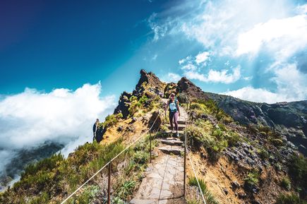 Person auf einem steinernen Bergpfad am Pico do Arieiro auf Madeira zwischen schroffen Felsformationen, Bergvegetation und vorbeiziehenden Wolken
