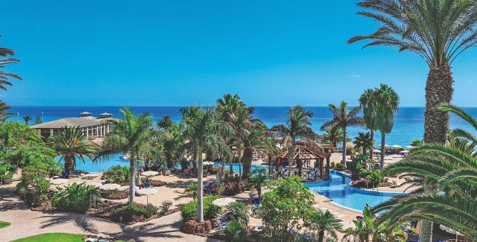 Weitläufige Garten- und Poolanlage des R2 Pájara Beach Hotel & Spa auf Fuerteventura mit zahlreichen Palmen, geschwungenen Pools und Blick auf das tiefblaue Meer unter klarem Himmel