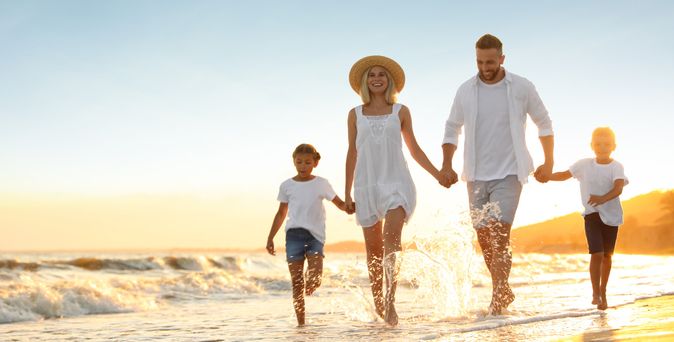 Familie mit zwei Kindern läuft am Strand bei Sonnenuntergang, alle tragen helle Kleidung