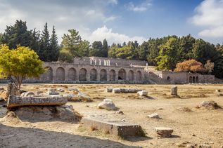 Ruinen des antiken Asklepieions auf Kos mit einer langen Arkadenreihe und verstreuten Steinen auf trockenem Boden vor bewaldetem Hintergrund