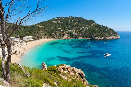Weite Bucht Cala de Sant Vicent mit Sandstrand, grünen Hügeln und Booten im Wasser
