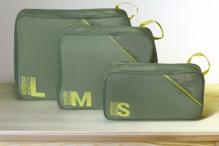 Drei grüne Packing Cubes von American Tourister in den Größen S, M und L auf einem hellen Holzboden vor hellem Hintergrund. Auf den Packing Cubes sind die Größenangaben sowie der Schriftzug „American Tourister Expand“ zu sehen.