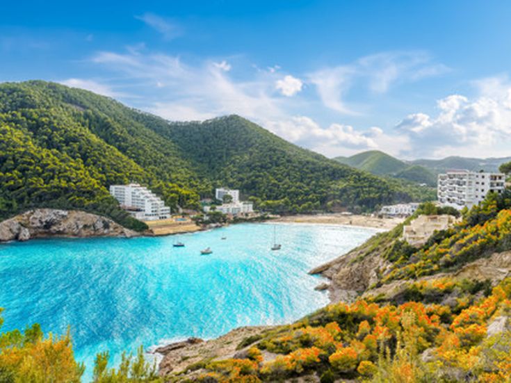 Blick auf die Bucht Cala Llonga auf Ibiza mit türkisblauem Meer, bewaldeten Hügeln, weißen Hotelgebäuden am Strand und Booten im Wasser
