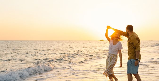 Mann und Frau tanzen lachend im seichten Wasser am Strand bei Sonnenuntergang