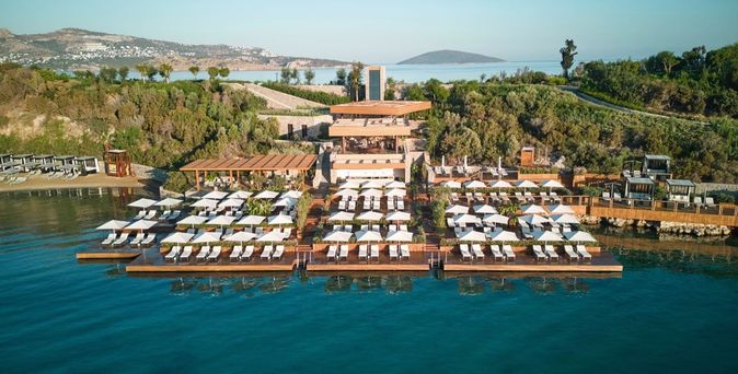 Holzstege des Maxx Royal Bodrum Resorts mit weißen Sonnenschirmen und Liegen am Wasser, umgeben von grünen Hügeln