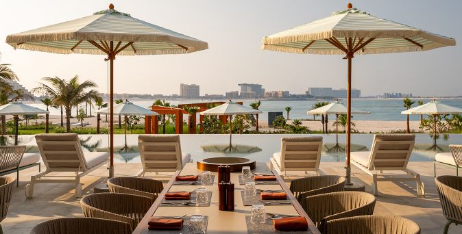 Mehrere gedeckte Tische mit Stühlen und Sonnenschirmen im Hotel SO/Ras Al Khaimah am Strand mit Blick auf das Meer und entfernte Gebäude.
