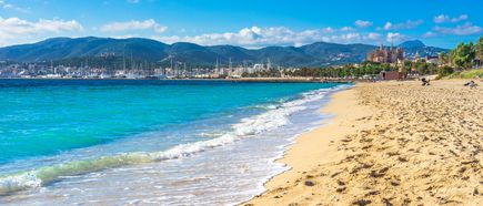 Sandstrand an der Playa de Palma auf Mallorca mit türkisblauem Meer, sanften Wellen, Palmenpromenade, Yachthafen und Bergkulisse im Hintergrund
