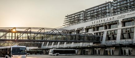 Flughafen Stuttgart Terminal 1 bei Sonnenaufgang mit Glasfassade, Fußgängerbrücke und Flughafenbussen vor dem Gebäude