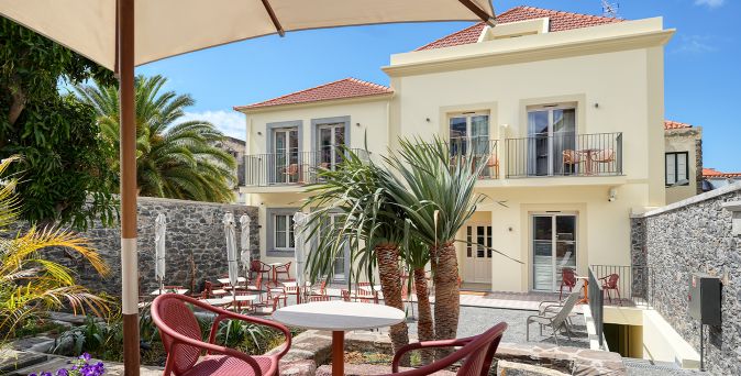 Außenansicht des Chapelaria Boutique Hotel mit hellem zweistöckigem Gebäude, Balkonen, tropischen Pflanzen und einem ruhigen Innenhof mit Tischen und roten Stühlen unter sonnigem Himmel