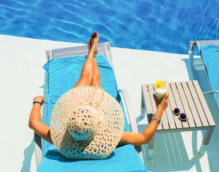 Frau mit großem Sonnenhut liegt auf blauem Handtuch auf Liegestuhl neben Pool, hält Cocktailglas, auf Tisch liegen Sonnenbrille und weitere Getränke.